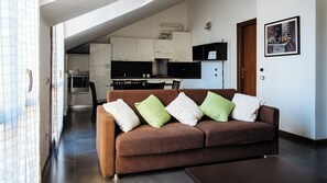 Een moderne woonkamer met een bruine bank, groene en witte kussens, en een keuken op de achtergrond.