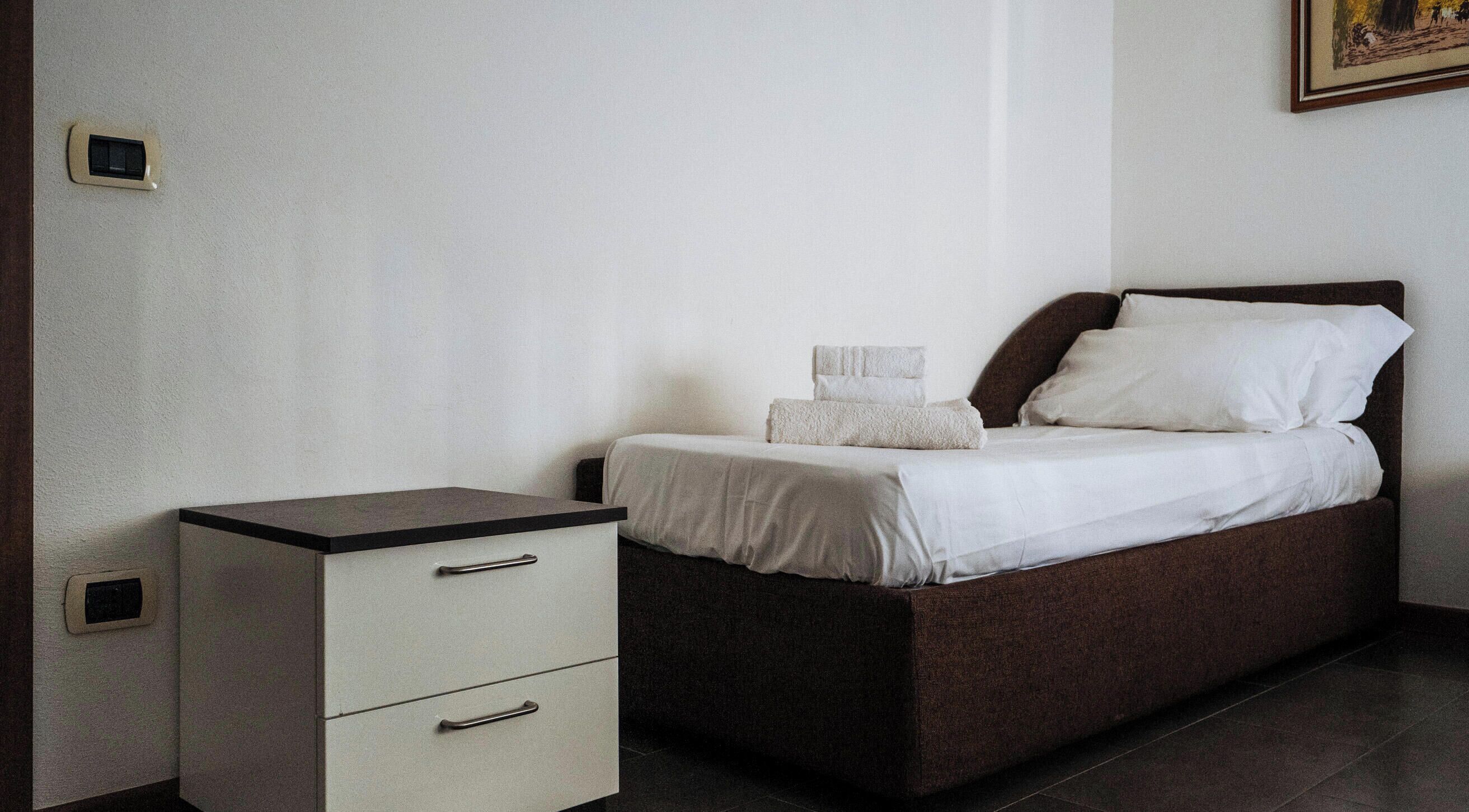 A single bed with a dark headboard and white bedding, a small bedside table with drawers, and a wall-mounted switch.
