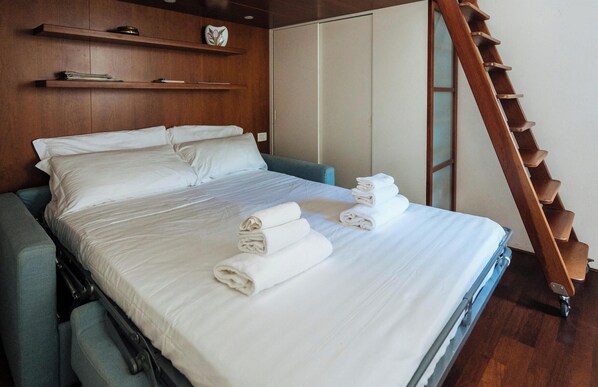 A neatly made bed with white linens, a wooden ladder, and a blue chair in a modern room with wooden paneling and a wardrobe. - Aurelia Harbour 103 (Andora)