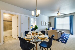 Un coin repas avec une table ronde dressée pour quatre personnes, un ventilateur de plafond et une vue sur une chambre à coucher avec un lit et une fenêtre avec des rideaux.