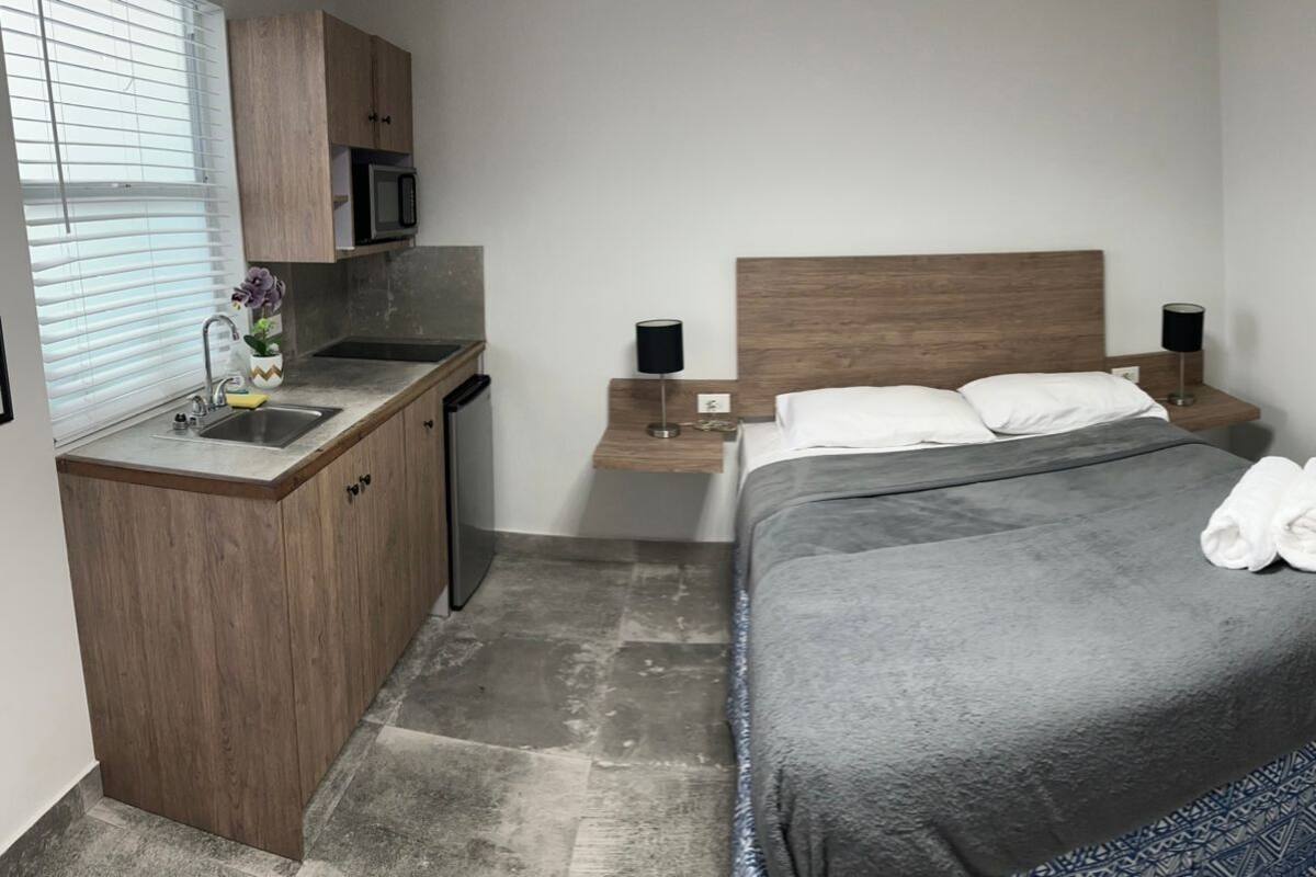 Ein Hotelzimmer mit einem Bett, einer kleinen Küchenzeile, einem Nachttisch mit Lampe und einer Tür.