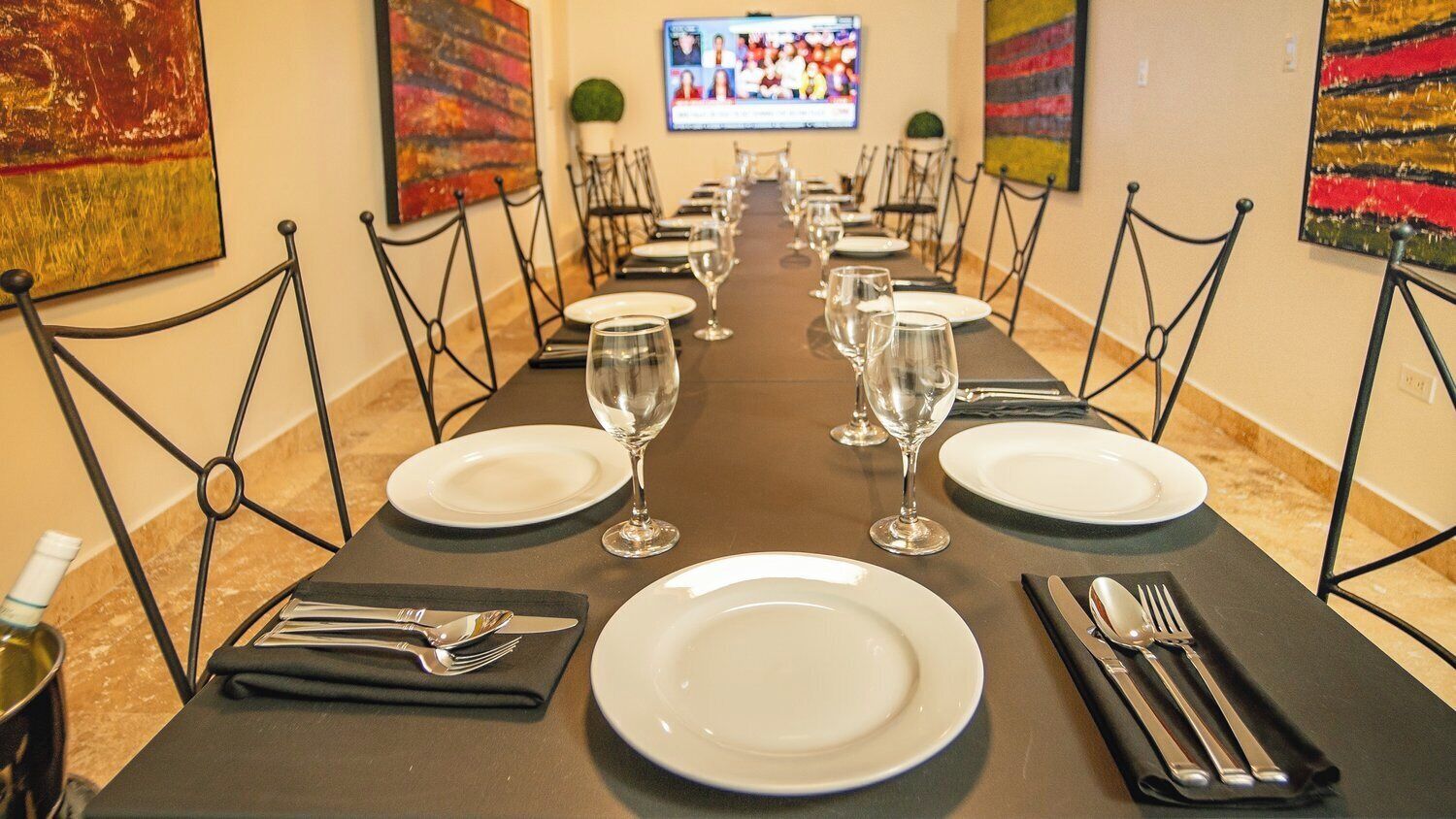Une longue table à manger, dressée avec de l’argenterie blanche, des verres à vin et des couverts, est entourée de chaises à structure métallique.