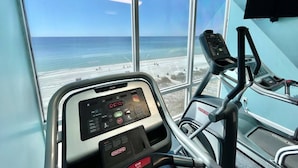 Een sportschool met een loopband en een digitaal scherm, met uitzicht op een strand.
