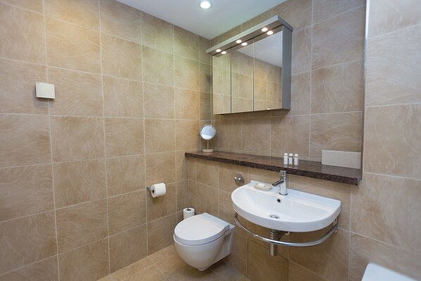 A modern bathroom with beige tiles, a white toilet, a sink with a mirror above, and a shelf with toiletries.