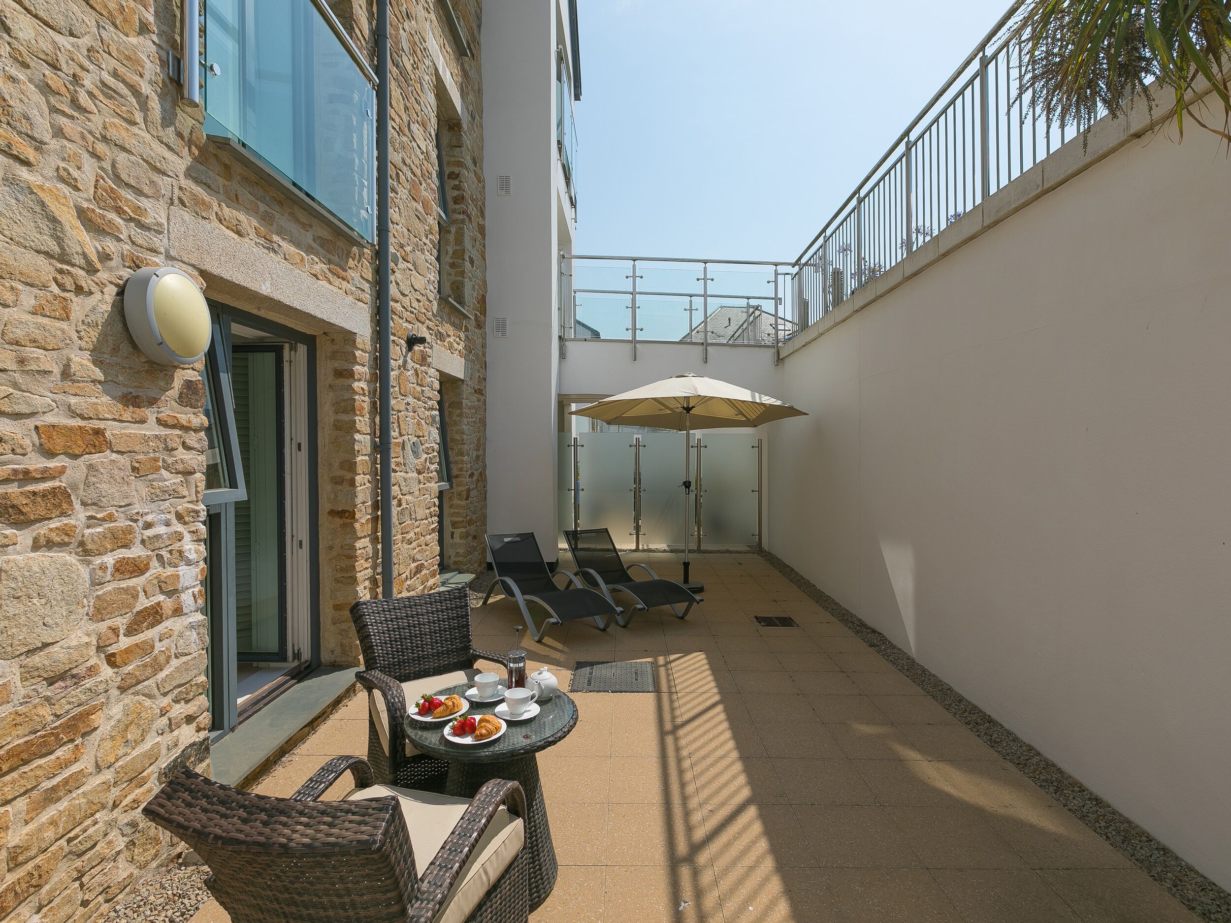 A stone building with a patio area featuring wicker chairs, a table with plates of food, and a large umbrella.