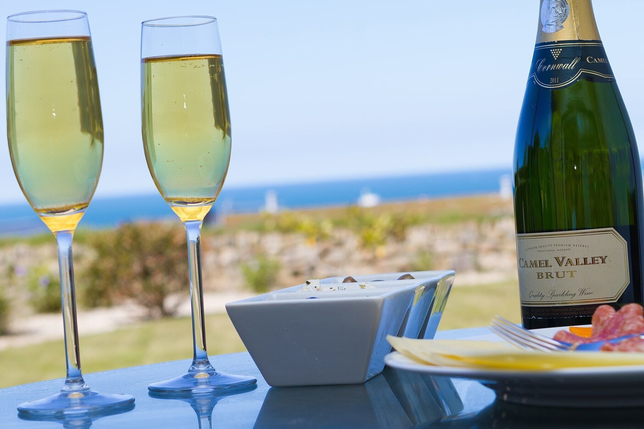Two glasses of white wine, a bottle of Camel Valley Brut, and a plate with food on a table with a view of the sea.