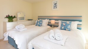 Une chambre d’hôtel avec deux lits, chacun doté de draps blancs et de serviettes pliées. On y trouve une table de chevet avec une lampe et un miroir. La chambre présente un aménagement simple et épuré, avec un mur de couleur claire et un tableau encadré au-dessus des lits.