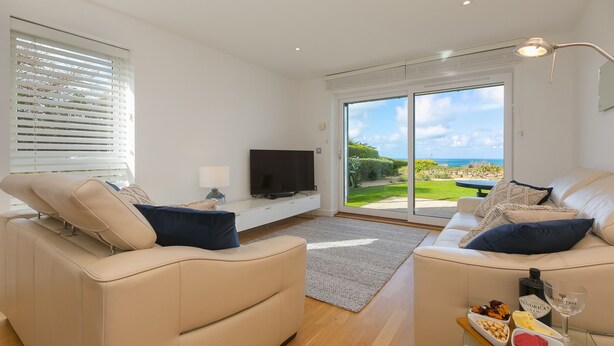 Un salon moderne avec deux canapés beige, un téléviseur à écran plat, une table basse en verre sur laquelle sont disposés des en-cas, et une grande porte-fenêtre coulissante donnant sur un balcon avec vue sur la mer.