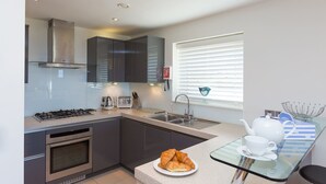 Une cuisine moderne équipée d’une cuisinière à gaz, d’un four et de placards en acier inoxydable. On y trouve une table en verre sur laquelle sont disposés une assiette de croissants et une théière.