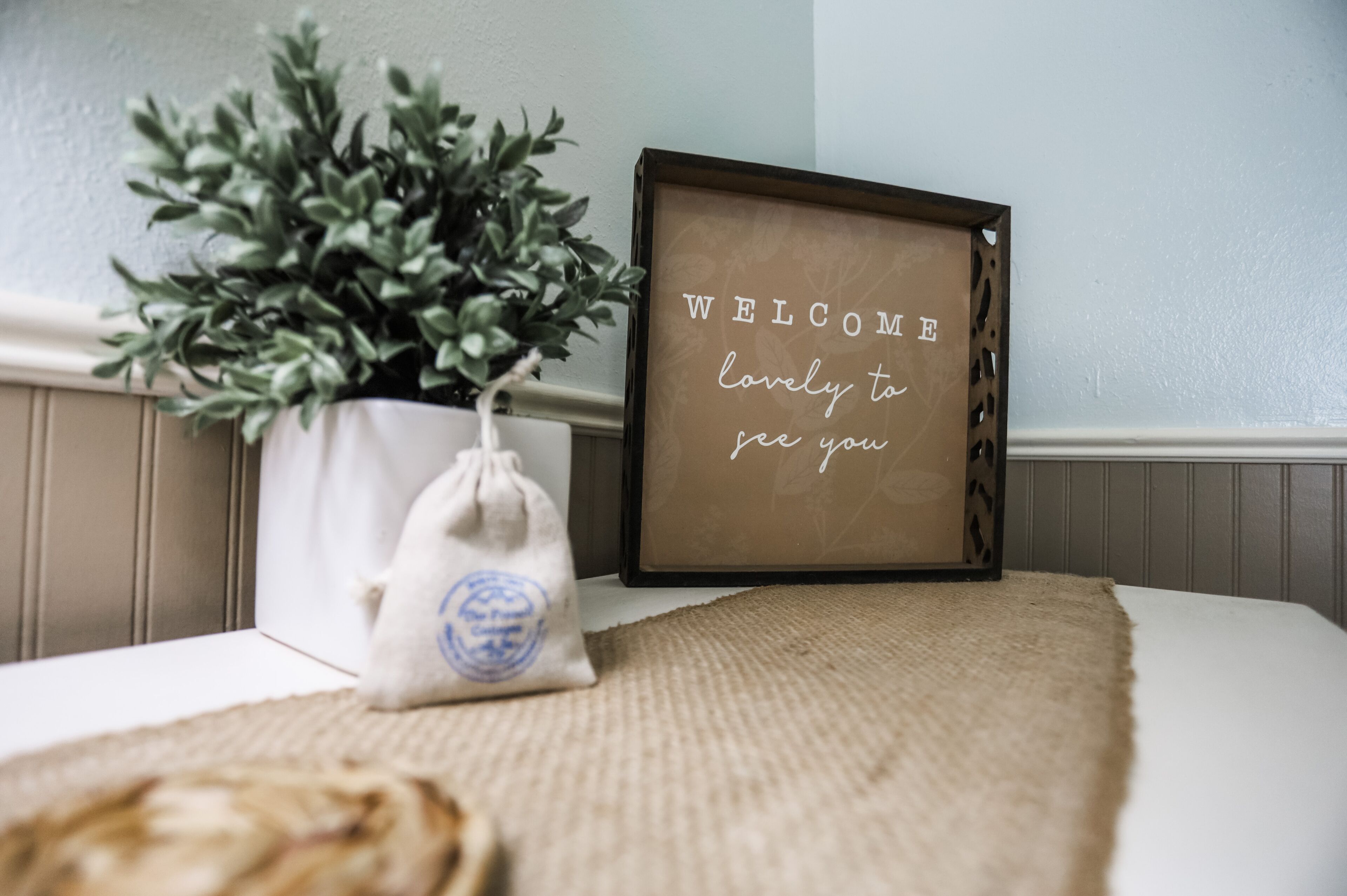 A framed welcome sign, a potted plant, and a small bag on a bed.