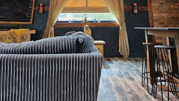 A rustic interior with exposed wooden beams, a fireplace, a large window with curtains, a sofa, and a bar area. - Tawitawoo Lodge Felmoor Park (Morpeth)