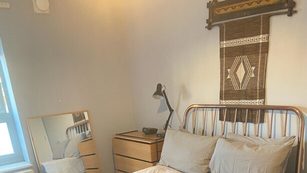 Un letto rifatto con una coperta grigia e due cuscini, una lampada da comodino, un comodino in legno e uno specchio sulla parete.