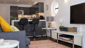 Una sala de estar moderna con un sofá, dos taburetes altos, una mesita y un televisor. Se ve una cocina al fondo con un microondas, horno y fregadero.