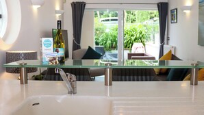 Un baño moderno con una encimera de vidrio, un lavabo y una botella de vino. Se aprecia una vista de una sala de estar con un sofá y una mesa de comedor.