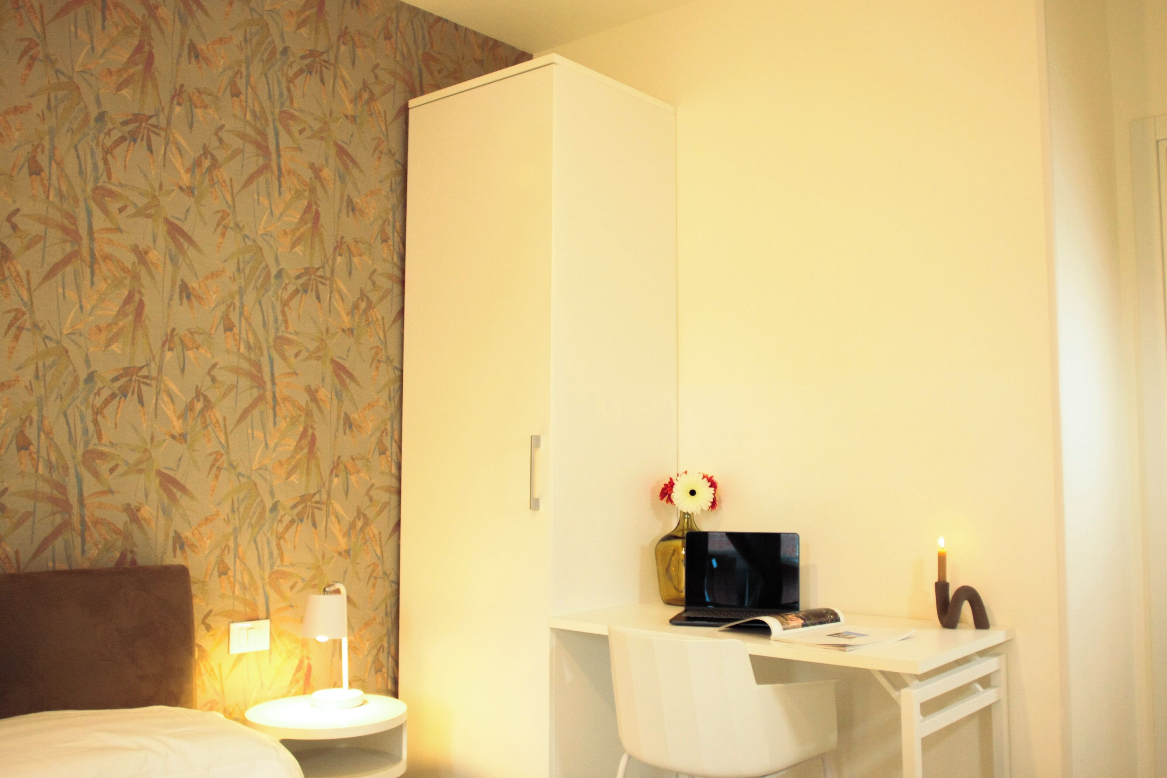 A hotel room with a bed, a desk with a laptop, a vase with flowers, a nightstand with a lamp, and a wardrobe.