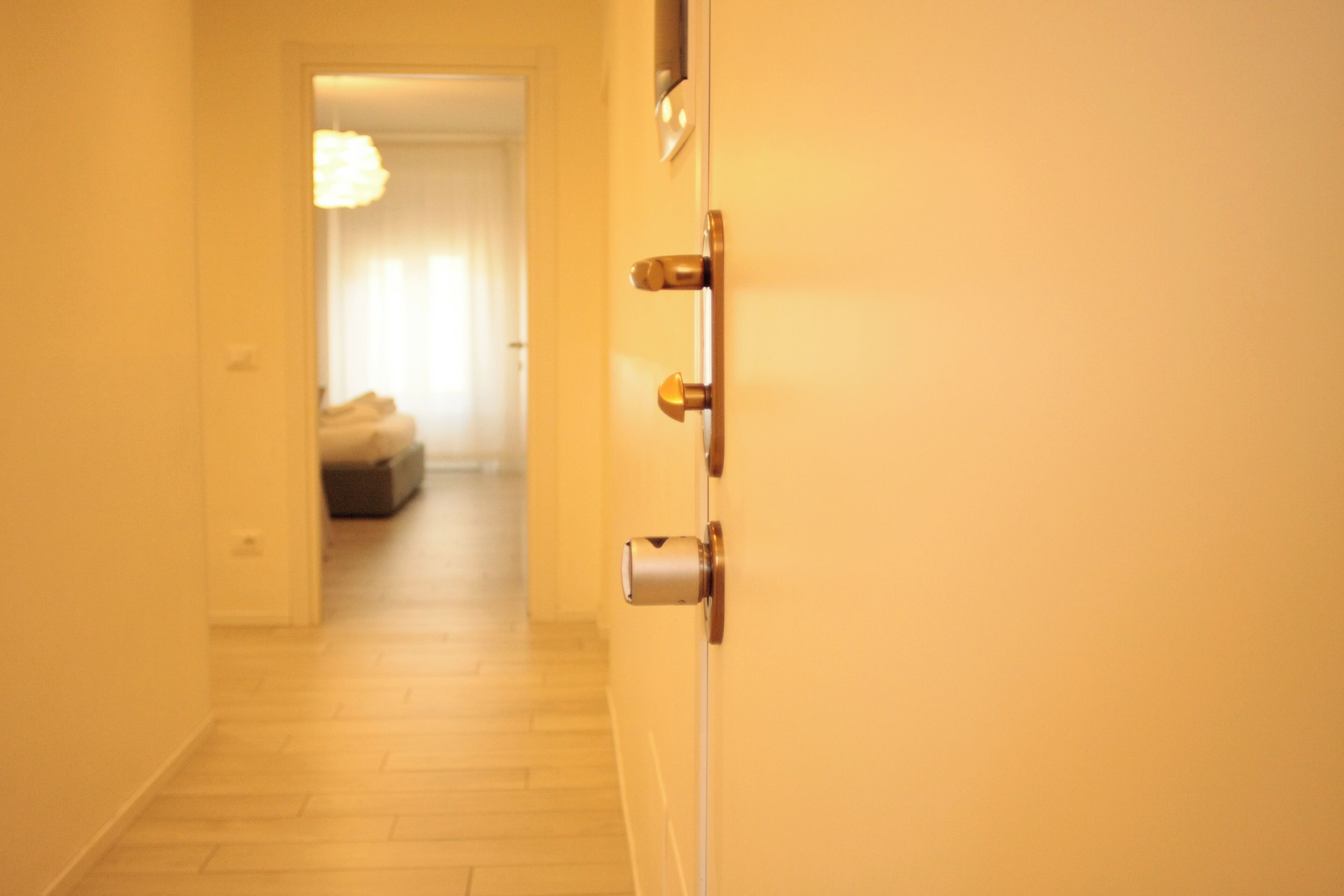 A hallway with a wooden floor leading to a room with a bed and a hanging light fixture.
