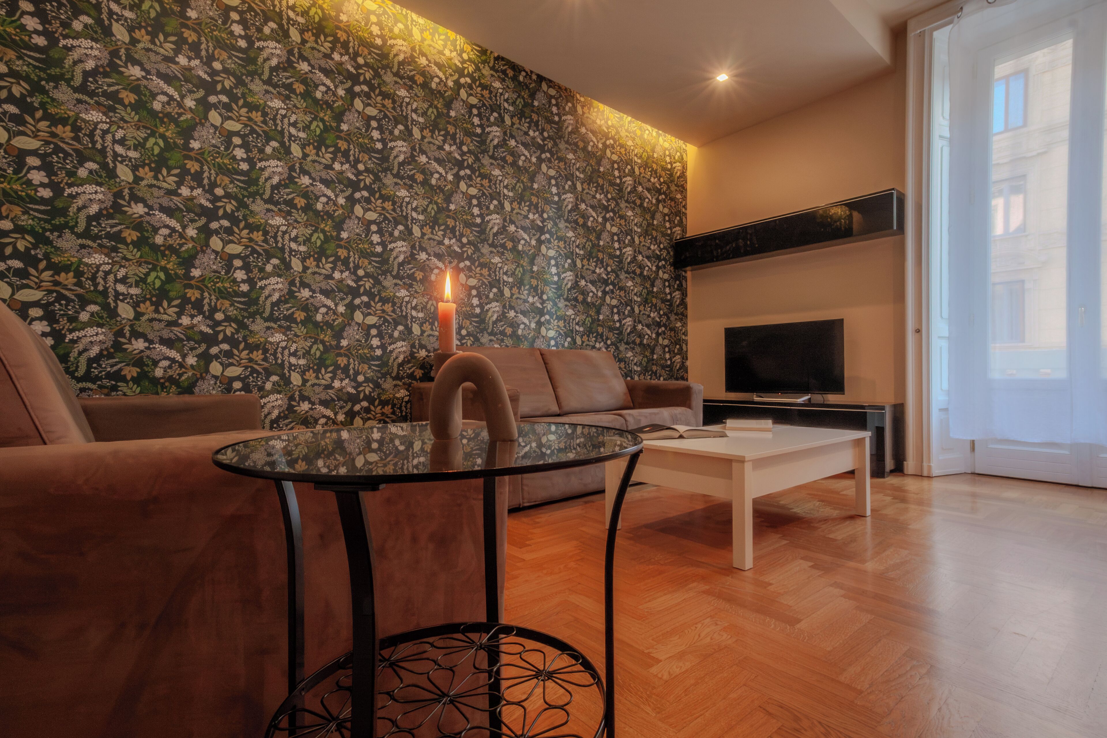 Een moderne woonkamer met bloemenbehang, een glazen salontafel, een bank en een televisie.