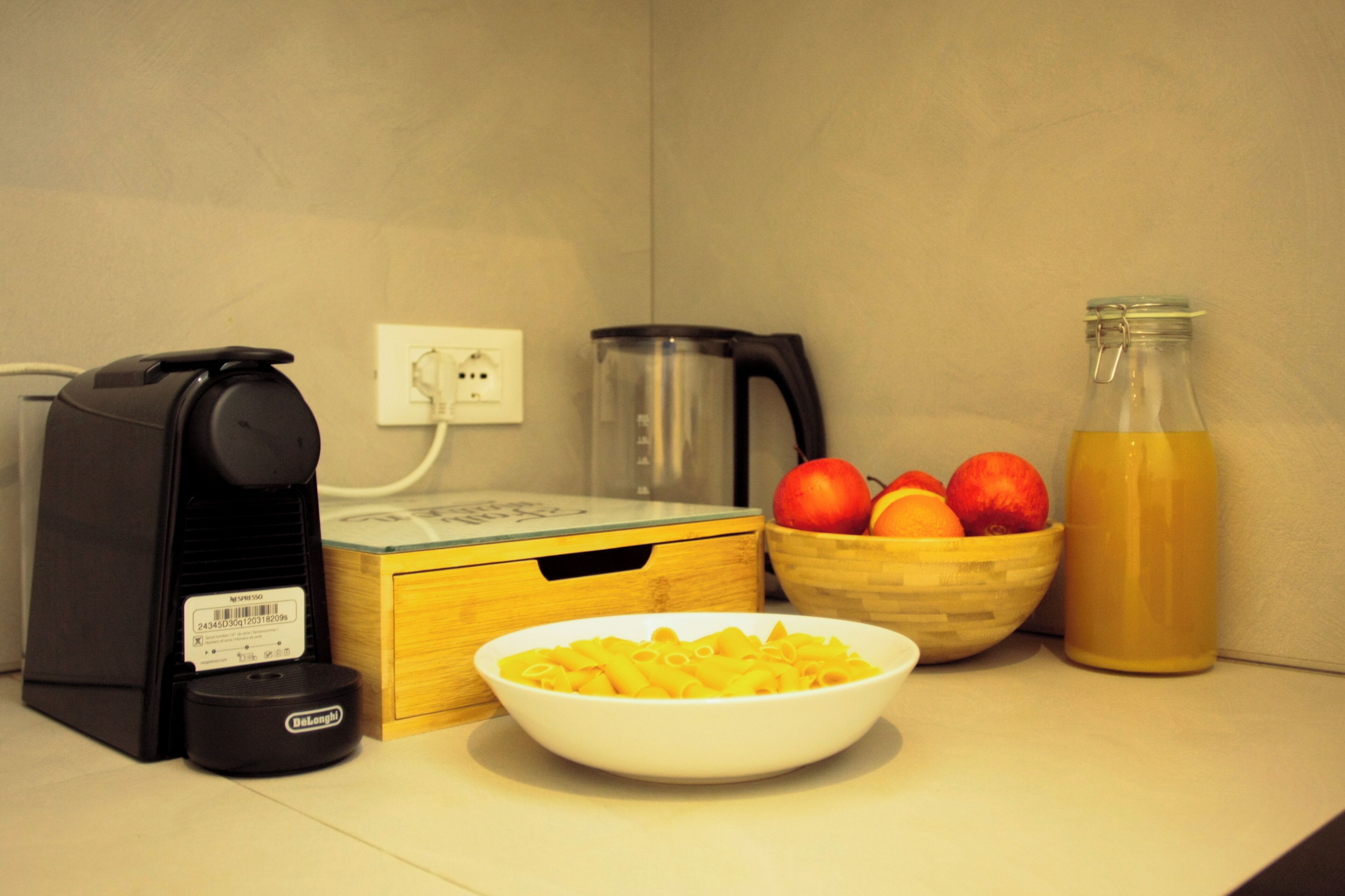 Un piano di lavoro in cucina con una macchina da caffè, un bollitore, una ciotola di pasta, un cesto di frutta e una bottiglia di succo.