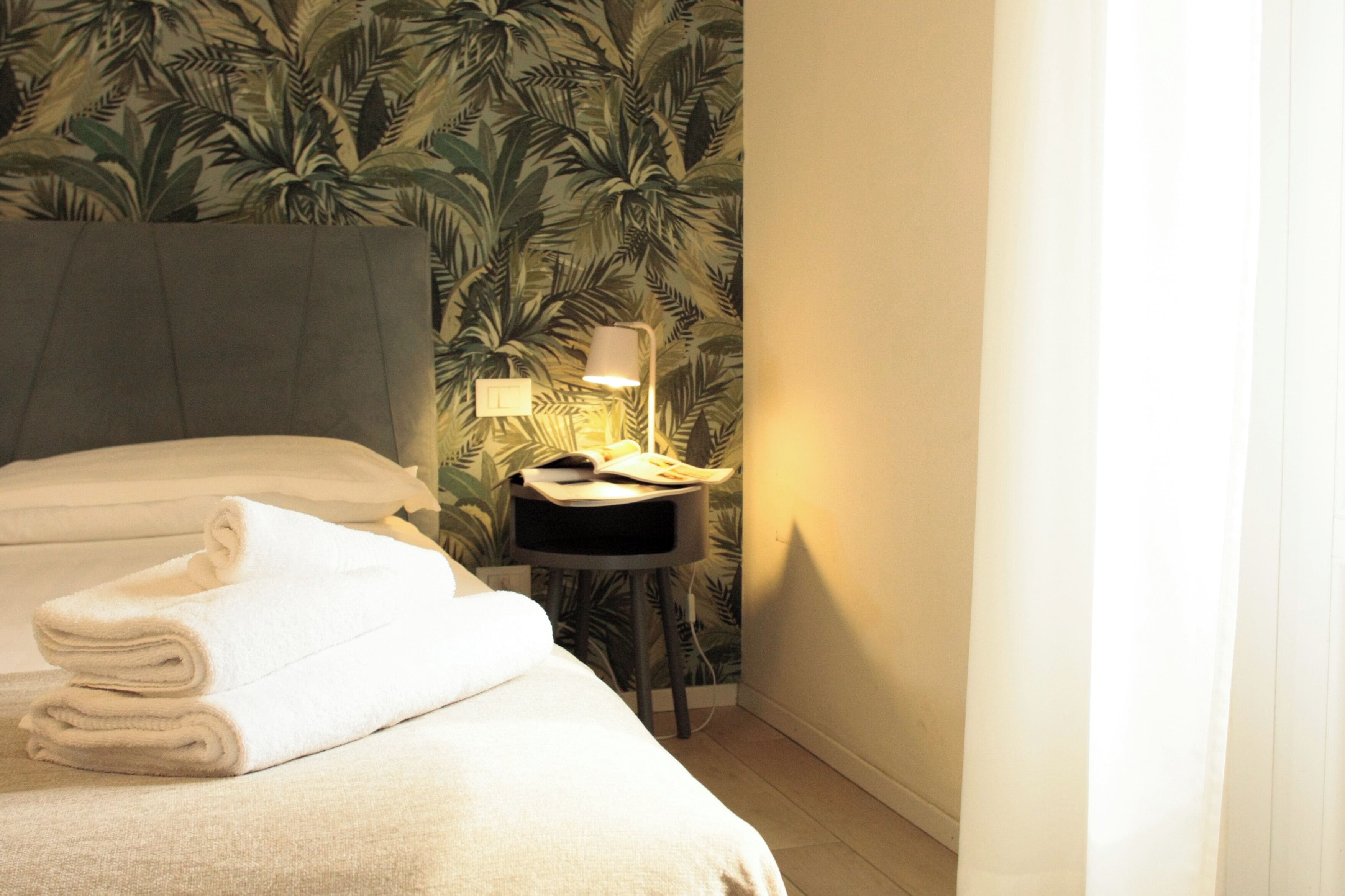 Un lit bien fait, avec des serviettes blanches pliées en rouleau, une lampe de chevet et une petite table sur laquelle se trouve une autre lampe.