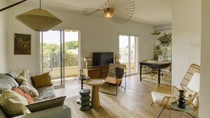 A spacious living area with a sofa, a coffee table, and a dining table with chairs. There is a television and a balcony with a view of greenery. - Very Nice 60m² With Balcony Near The Beaches! (Six-Fours-les-Plages)