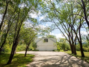 A white barn with a gabled roof and a central window, surrounded by trees and a gravel driveway.