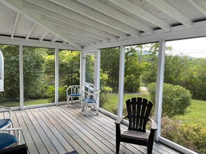 A covered outdoor deck with wooden flooring, white railings, and large windows offering a view of a lush garden and trees.