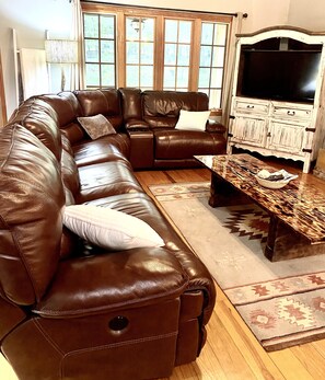 Ein braunes, ledernes Ecksofa mit passendem Hocker, ein Couchtisch aus Holz mit einer Schale und ein weißes Schränkchen mit einem Fernseher.