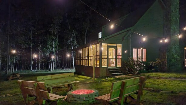 A cozy outdoor seating area with wooden benches and a fire pit, surrounded by a green house with large windows and string lights.