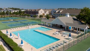 En swimmingpool med liggestole og parasoller, omgivet af tennisbaner og boligbygninger.