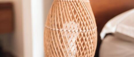 A wooden bedside table with a woven lamp and a white pillow on a bed.
