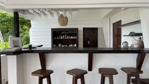 A kitchen with a bar area, wooden stools, a glassware rack, and a shelf with kitchenware. - Le M Hotel Marie Galante (Capesterre-de-Marie-Galante)