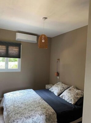 A bedroom with a bed, a window with blinds, a wall-mounted air conditioner, and a hanging light fixture.