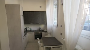 A compact kitchen with a dining table and chairs, a refrigerator, dark cabinets, and a window with curtains.