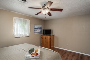A bedroom with a bed, a ceiling fan, a wooden cabinet, a television, and a window with sheer curtains.