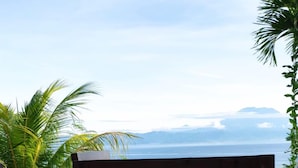A table with a plate of food, a glass of orange juice, and a view of the ocean and mountains. - Gara Gara Sea View (Penida Island)