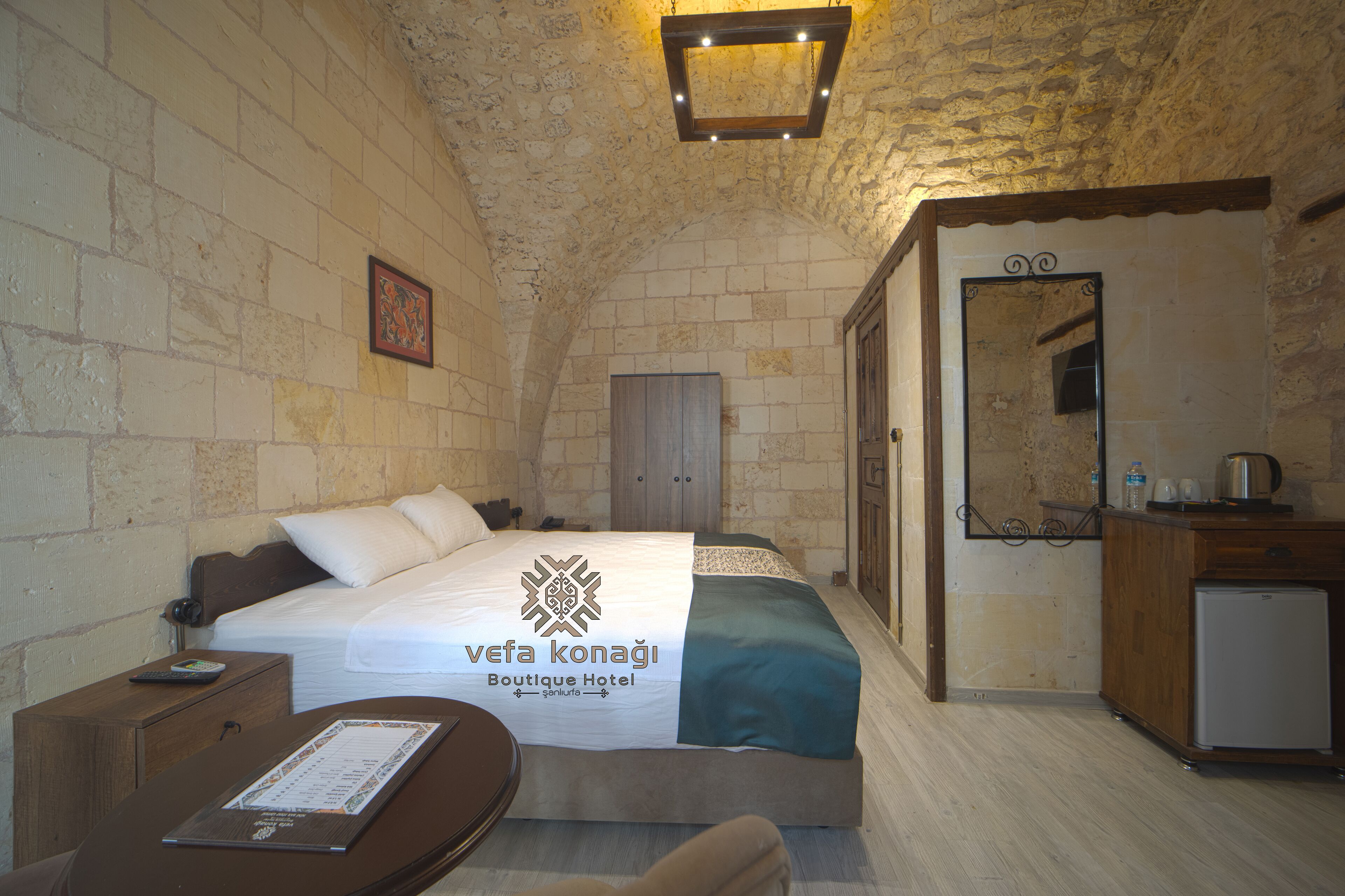 Ein Hotelzimmer mit einer Steinwand, einem Bett mit weißer und türkiser Bettdecke, einem Nachttisch aus Holz, einem kleinen Schreibtisch mit Stuhl, einer Schreibtischlampe, einem Spiegel und einem Kühlschrank.