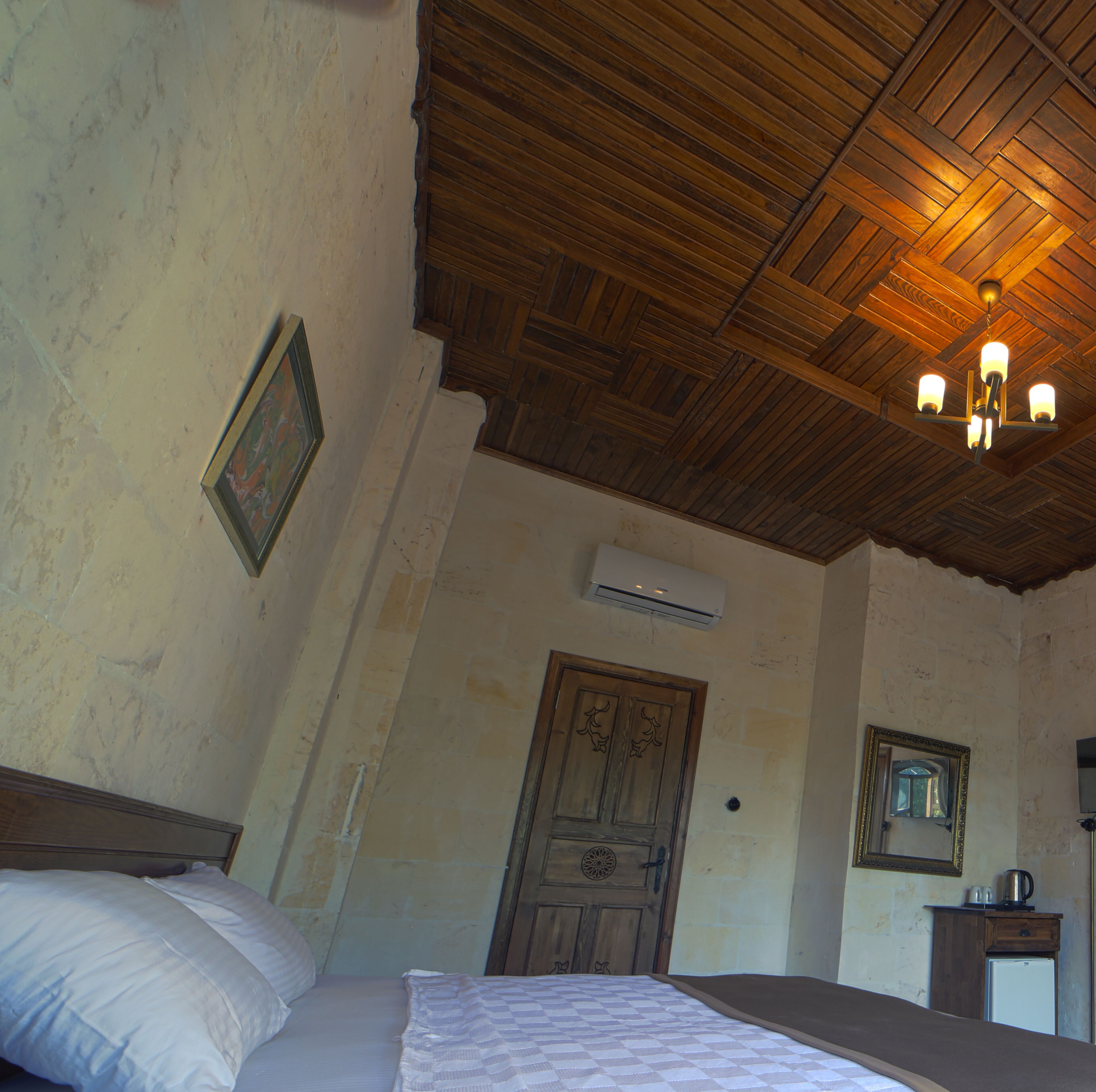 A bedroom with a wooden ceiling, a bed with white bedding, a wooden door, a small cabinet, and a wall-mounted air conditioner.