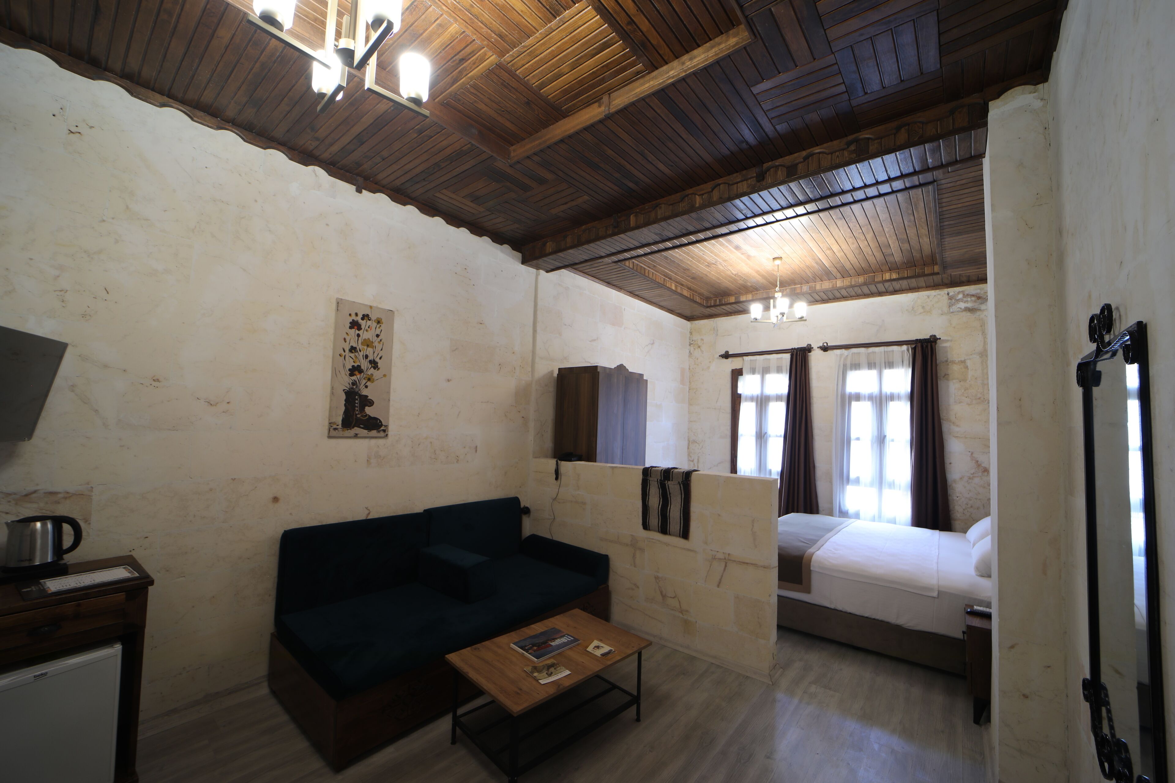 A room with a wooden ceiling, a dark-colored sofa, a small table with books, a bed with a white mattress, and a window with curtains.