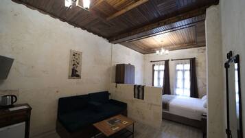 A room with a wooden ceiling, a dark-colored sofa, a small table with books, a bed with a white mattress, and a window with curtains.