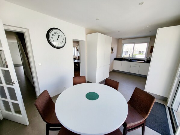 A compact kitchen with white cabinets, a dining area with a round table and brown chairs, and a wall clock.