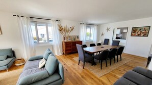 Ein modernes Wohnzimmer mit einer Couch, einem Esstisch und Stühlen. Große Fenster mit Vorhängen, eine Heizung und eine Vitrine mit Dekoration.