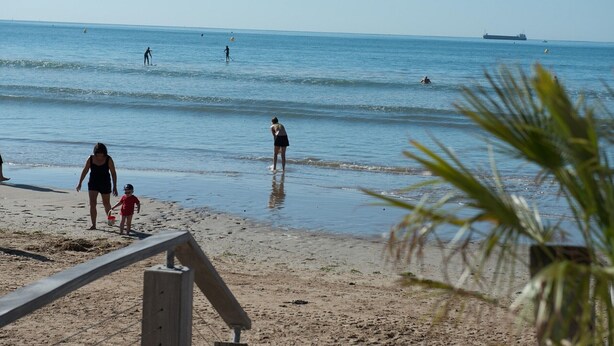 Ein Strand mit einer Holzpromenade, die zum Wasser führt, Menschen schwimmen und spazieren am Ufer entlang.