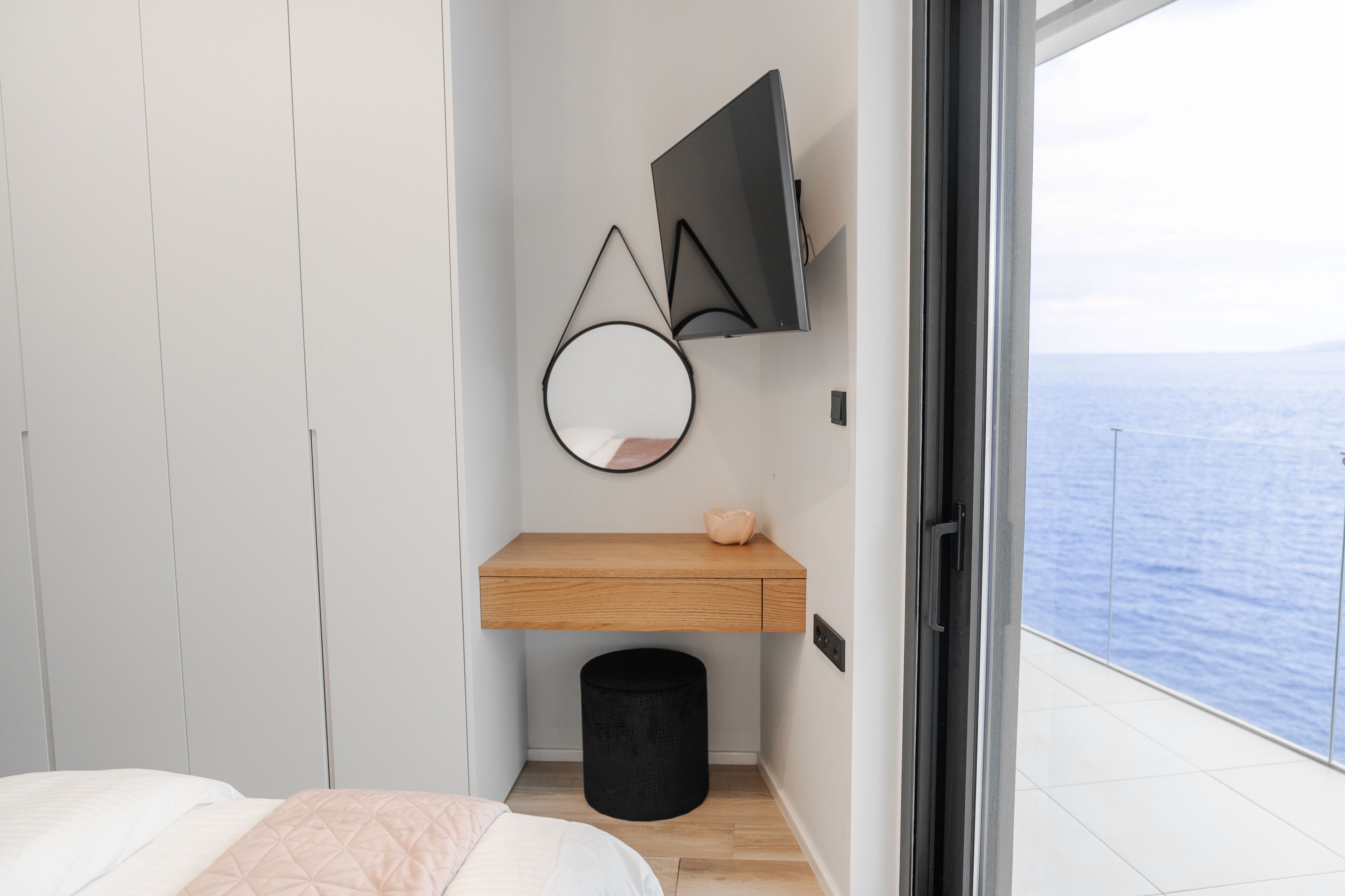 Ein modernes Schlafzimmer mit Bett, einem Holfnachtkästchen, einer schwarzen Mülltonne, einem runden Spiegel und einem an der Wand befestigten Fernseher.