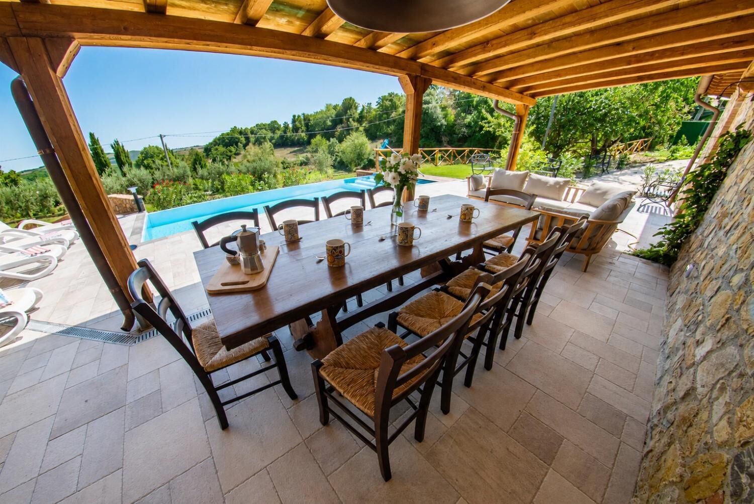 An outdoor dining area with a pool view, wooden pergola, and stone walls.