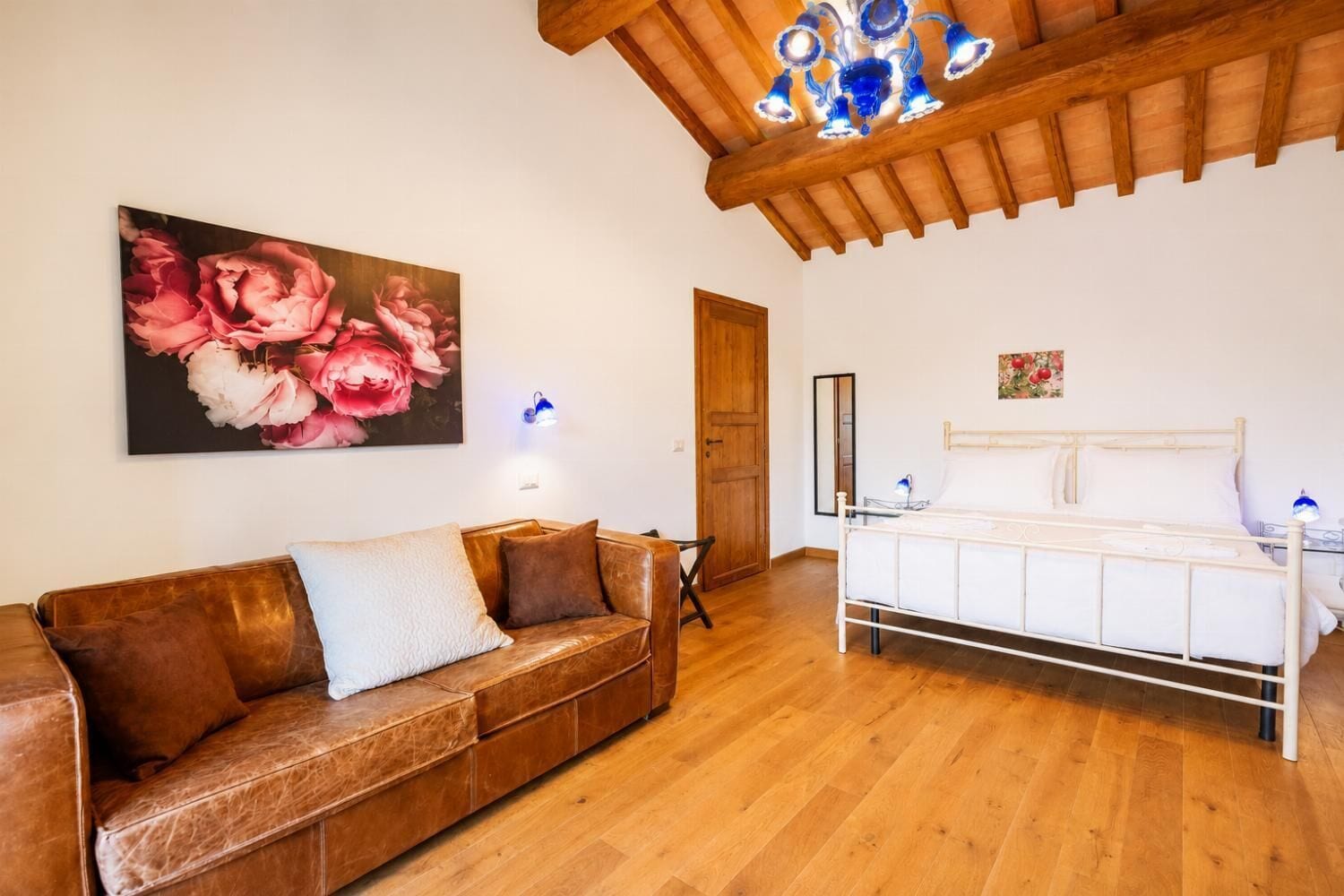 Une pièce avec un plancher en bois, un canapé en cuir marron, un lit blanc, un plafond en bois et un grand tableau représentant des motifs floraux.