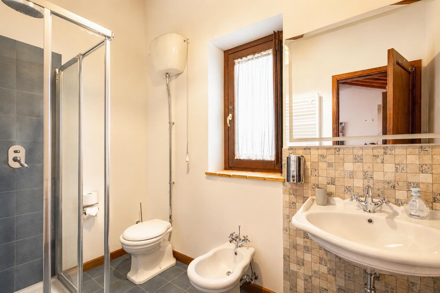 Un baño con ducha acristalada, inodoro, bidé, lavabo con espejo y ventana con cortinas.
