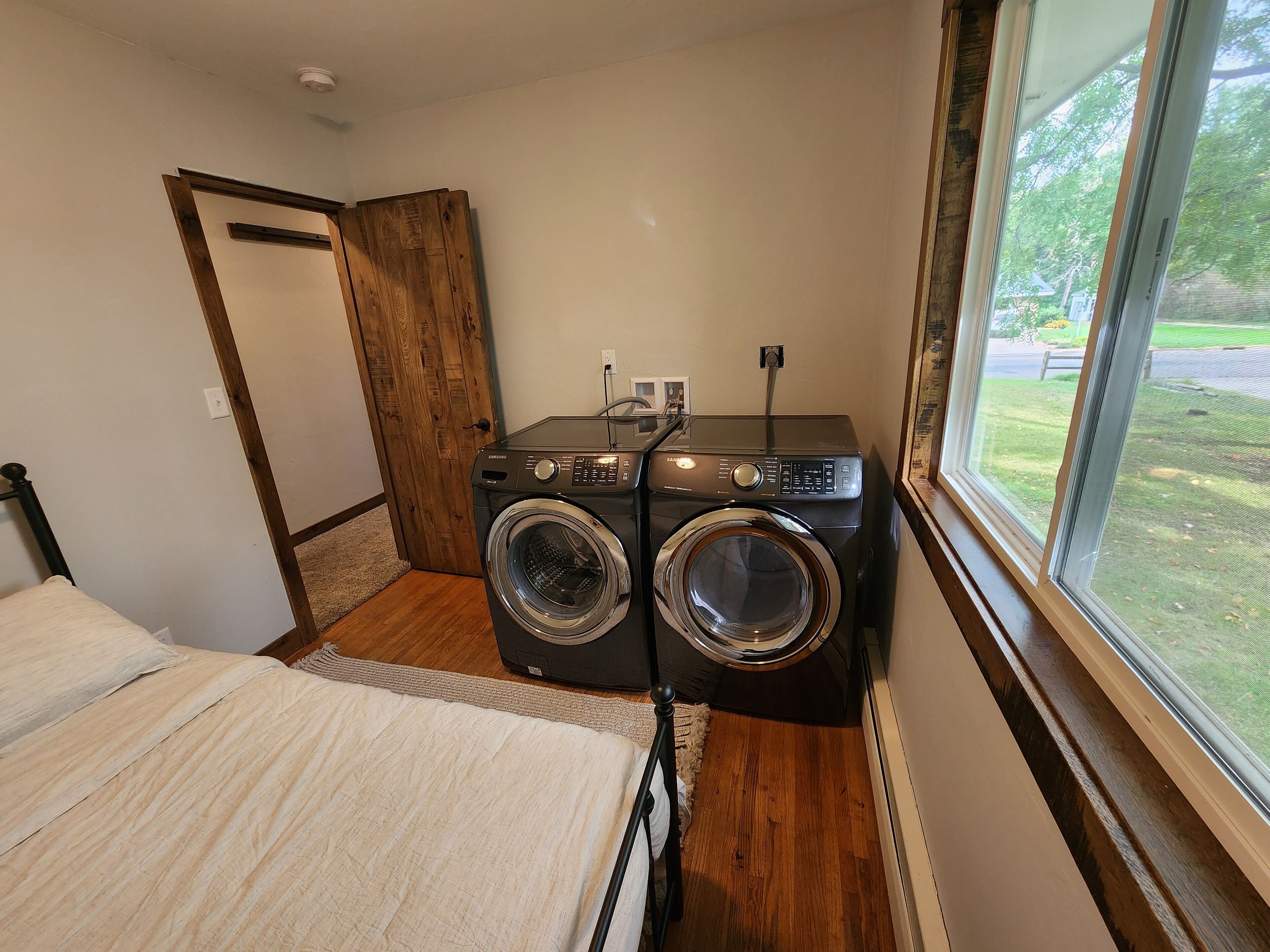 A compact laundry room with two front-loading washing machines, a wooden door, and a large window offering a view of the outdoors.