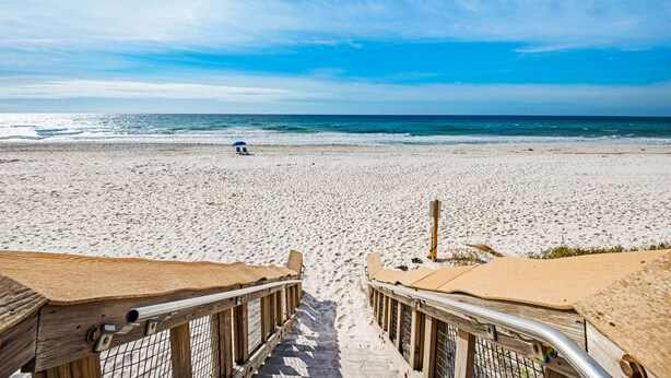 A beach with a wooden walkway leading to the water, a person with an umbrella, and a clear sky. - 🌞Stunning Balcony Beachfront Gulf View | Pool | 2BR Sundunes112🌞 (Navarre)