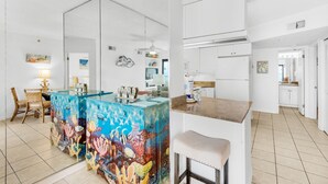A modern kitchen with white cabinetry, a blue painted island with coral designs, and a small bar area with a stool. - 🌞Stunning Balcony Beachfront Gulf View | Pool | 2BR Sundunes112🌞 (Navarre)