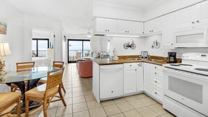 A modern kitchen with white cabinets, a dining area with a glass table, wicker chairs, and a view to the outside. - 🌞Stunning Balcony Beachfront Gulf View | Pool | 2BR Sundunes112🌞 (Navarre)